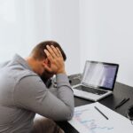 Een man die met zijn hoofd in zijn handen aan een bureau zit met een laptop en allemaal papieren